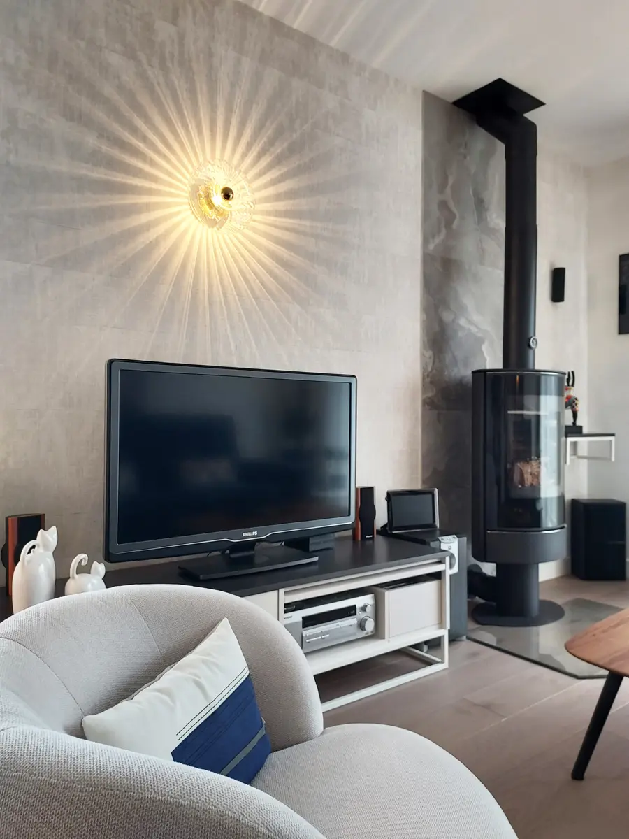 Espace télévision, applique soleil dorée, poêle à bois cylindrique, fauteuil beige confortable